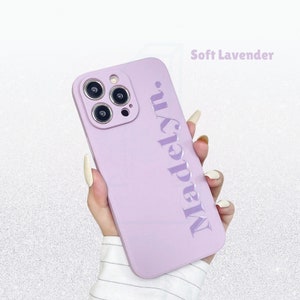 May include: A soft lavender phone case with the name "Madelyn" embossed vertically. The case has a smooth finish and is designed for a smartphone with multiple camera lenses. The text "Soft Lavender" is in the upper right corner.