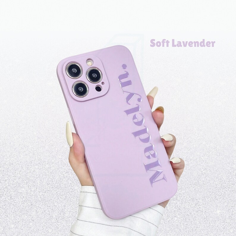 May include: A soft lavender phone case with the name "Madelyn" embossed vertically. The case has a smooth finish and is designed for a smartphone with multiple camera lenses. The text "Soft Lavender" is in the upper right corner.