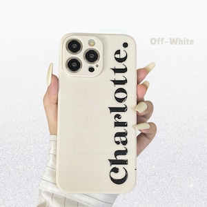 May include: Off-white phone case featuring the name "Charlotte" printed vertically in a bold, black font. The case has a camera cutout and is held in a hand.