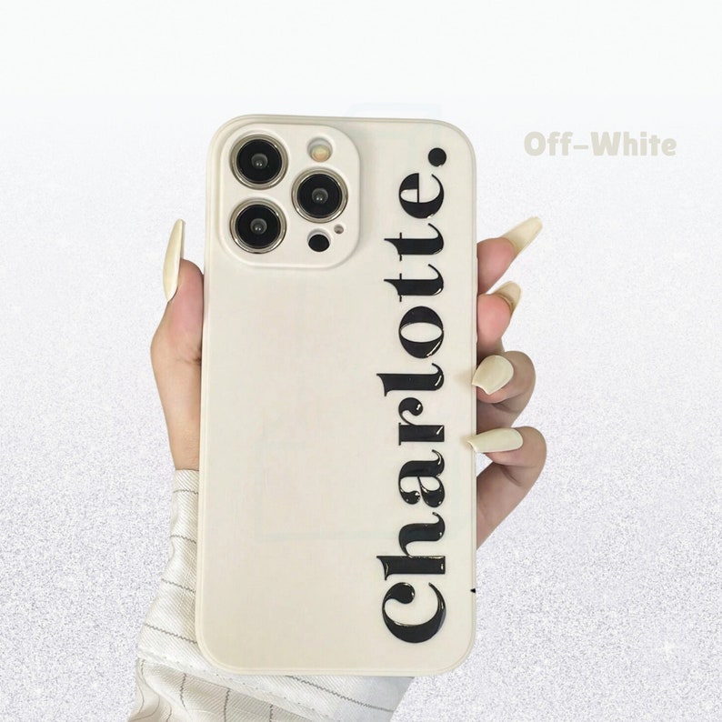 May include: Off-white phone case featuring the name "Charlotte" printed vertically in a bold, black font. The case has a camera cutout and is held in a hand.