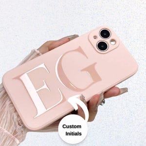 May include: A light pink phone case with the letters "EG" in a glossy, raised design. The case has a smooth finish and features a camera module. The text "Custom Initials" is in a white circle.