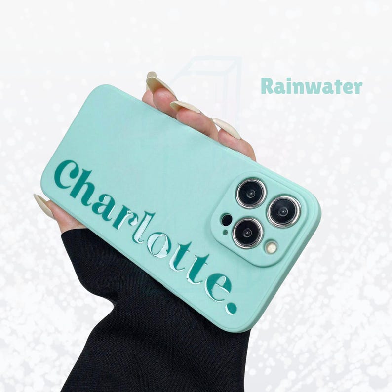 May include: A light teal phone case with the name "Charlotte" in a cut-out font. The case has a smooth finish and features a camera module with three lenses. The word "Rainwater" is also visible.