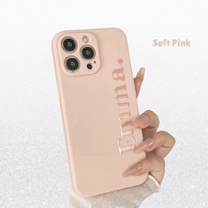 May include: A soft pink phone case with the name "Emma" embossed vertically. The case has a smooth finish and features a camera cutout. The text "Soft Pink" is displayed in the upper right corner.