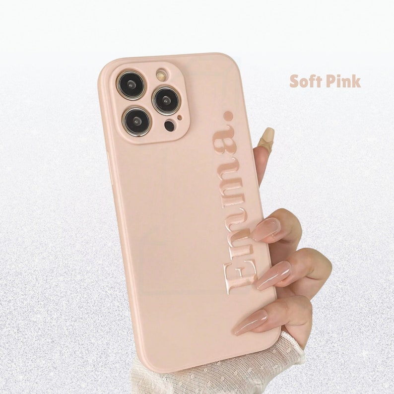 May include: A pale pink phone case with the name "Emma" embossed vertically. The case has a smooth finish and a camera cutout. The text "Soft Pink" is displayed in the upper right corner.