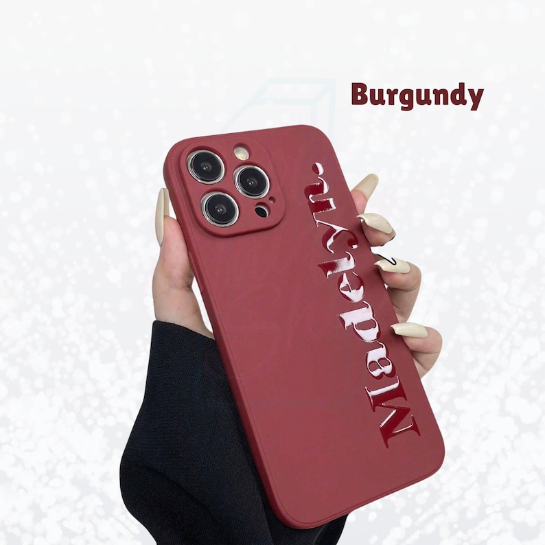 May include: Burgundy phone case with the name "Marilyn" in a glossy, raised font. The case has a matte finish and a camera cutout for multiple lenses.