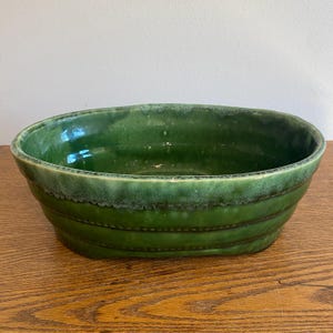 May include: An oval-shaped ceramic bowl with a glossy, deep forest green glaze. The exterior features horizontal ribbed detailing along the sides, while the rim displays a lighter, mottled green finish with a smooth, rounded edge and a wide opening.