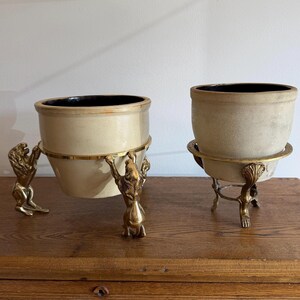May include: Two ceramic pots with dark interiors, each held by a brass stand. The left stand features three lion figures as legs, while the right stand has ornate, clawed feet. Both pots have a smooth, light-colored exterior finish and a wide rim.