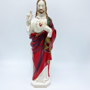 May include: A ceramic statue of Jesus Christ, featuring a white robe, a red cloak, and a golden heart emblem. The statue has brown hair and a beard, with one hand raised in blessing. The base is a hexagonal shape.
