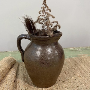May include: A brown stoneware pitcher with a handle, holding dried flowers and a thistle. The pitcher has a textured surface and a slightly flared rim. The background includes a neutral wall and a textured fabric.