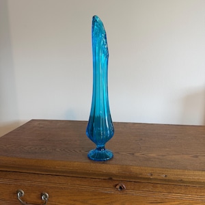 May include: A tall, slender, vibrant turquoise blue glass vase featuring a vertical ribbed texture. The vase has a flared, asymmetrical opening at the top, a narrow neck, a bulbous lower body, and a circular pedestal base made of matching glass.