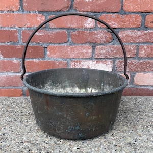 May include: A weathered, dark metal bucket with a rounded basin and a thin, arched handle attached to the sides. The exterior surface shows signs of oxidation and discoloration, featuring a mottled brown and grey patina throughout the metal.