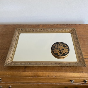 May include: Rectangular gold-framed mirror with ornate detailing. A small, round black and gold decorative box with a floral design sits on the mirror's surface. The mirror and box are on a wooden surface.