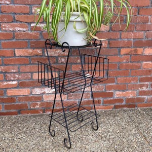 May include: A black metal plant stand with three tiers, featuring a top shelf, a middle basket-style shelf, and a bottom shelf. The structure is supported by thin metal legs with curled feet and decorative scrollwork accents at the top.
