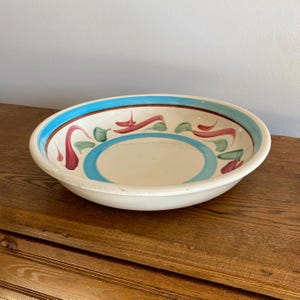 May include: Oval ceramic bowl with a cream-colored interior and a wide, light blue rim. The bowl features a decorative pattern of red, green, and blue brushstrokes. The bowl is sitting on a wooden surface.