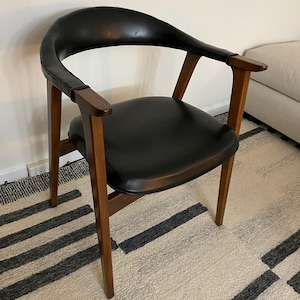 May include: A mid-century modern armchair with a curved black leather back and seat. The chair features a wooden frame with armrests and legs. The chair is set on a patterned rug.