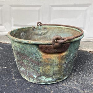 May include: A weathered copper bucket with a rounded body and a sturdy handle. The bucket has a patina of green and brown hues, with visible signs of age and use. The handle is made of a thick, rusty metal, and is attached to the bucket with rivets.