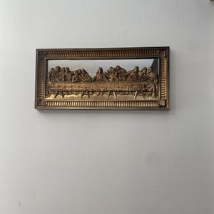May include: A rectangular wall plaque featuring a three-dimensional gold-toned relief of The Last Supper. The scene depicts thirteen figures seated at a long table, encased within an ornate, textured gold frame with decorative corner details.