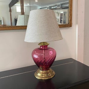 May include: A table lamp featuring a translucent, deep pink, heart-shaped glass base with a polished brass pedestal and neck. The lamp is topped with a tapered, light beige, textured fabric lampshade that provides a neutral contrast to the glass.
