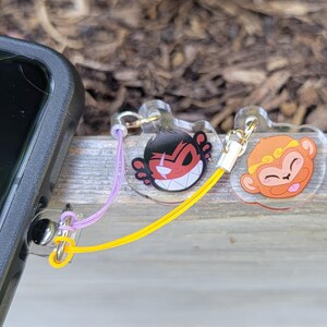 May include: Two clear acrylic phone charms with cartoon monkey faces. One charm has a red monkey face with a mischievous grin and the other has a yellow monkey face with a tongue sticking out. The charms are attached to a phone case with a purple and yellow string.