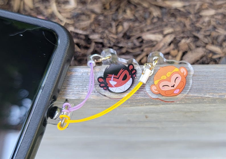 May include: Two clear acrylic phone charms with cartoon monkey faces. One charm has a red monkey face with a mischievous grin and the other has a yellow monkey face with a tongue sticking out. The charms are attached to a phone case with a purple and yellow string.