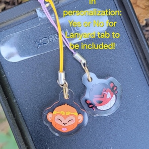 May include: Two colorful phone charms with cartoon monkey and demon faces.  The charms are attached to a black phone case with a lanyard tab.  In personalization: Yes or No for Lanyard tab to be included!