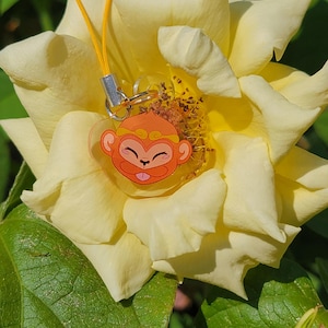 May include: A yellow rose with a small orange monkey charm attached to it. The charm is made of clear plastic and has a gold string attached to it.