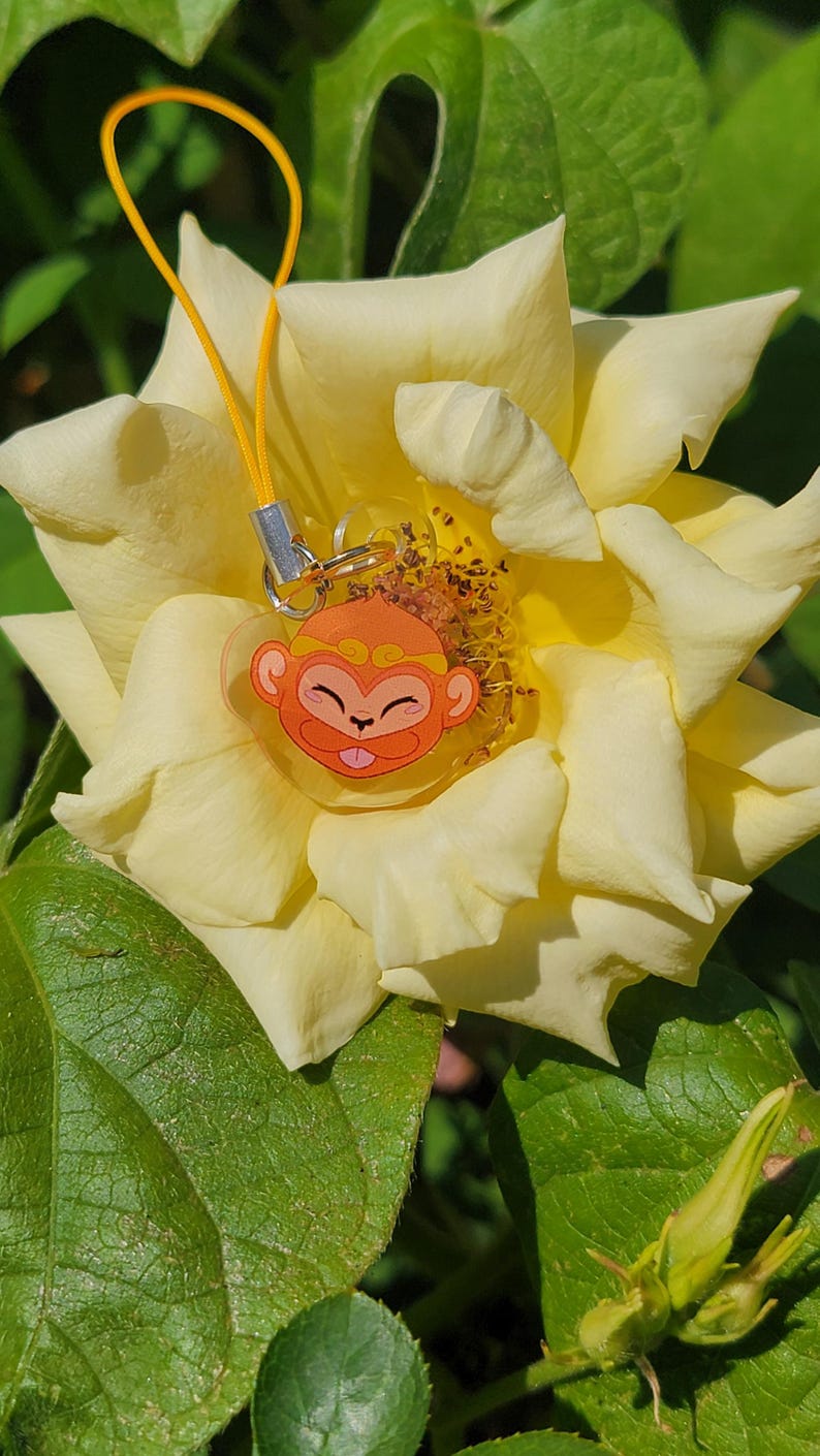 May include: A yellow rose with a small orange monkey charm attached to it. The charm is made of clear plastic and has a gold string attached to it.