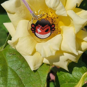 May include: A red and black cartoon monkey face charm with a purple lanyard hanging from a yellow rose.