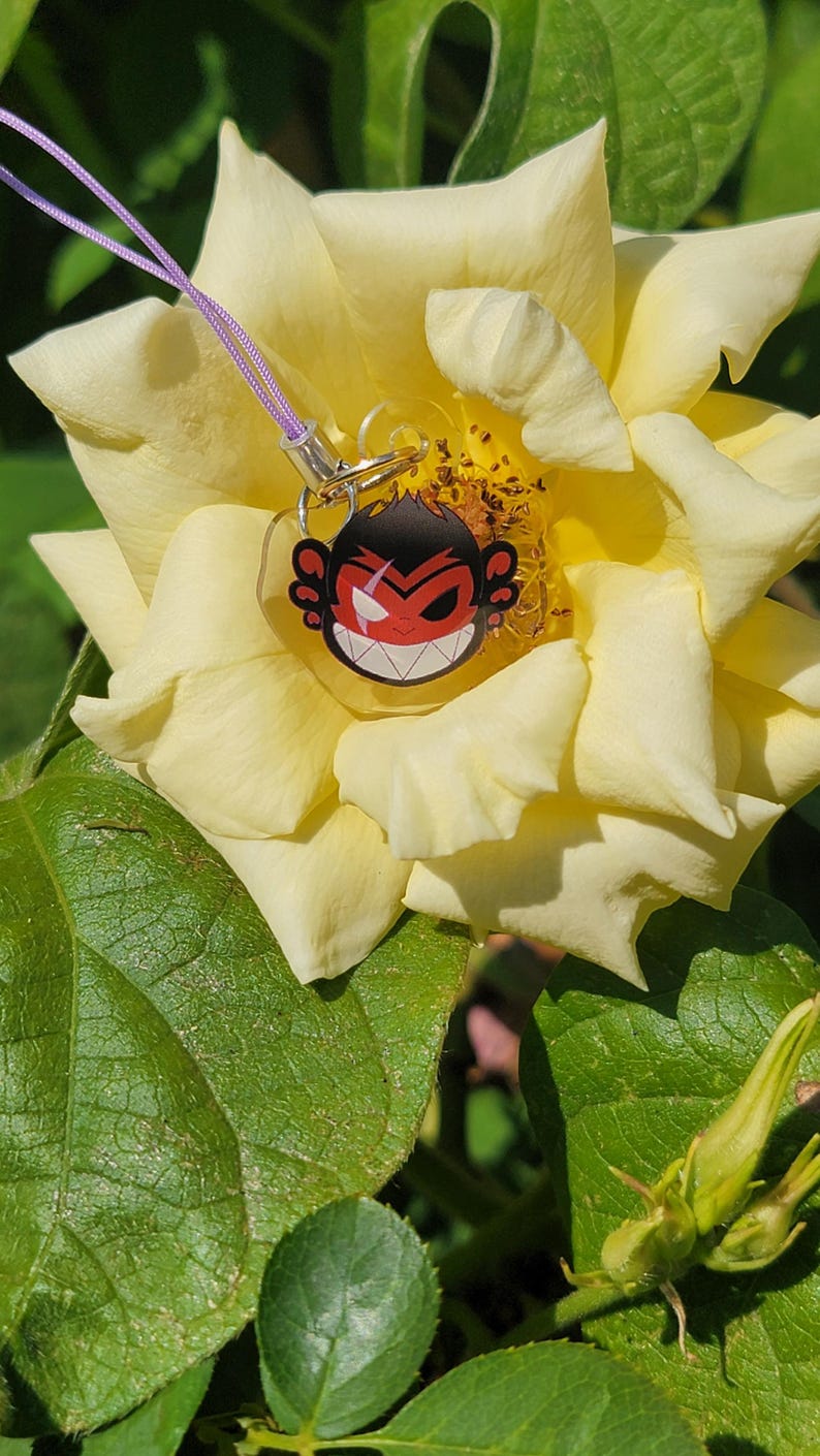May include: A red and black cartoon monkey face charm with a purple lanyard hanging from a yellow rose.