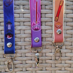 May include: Three colorful lanyards with metal clasps. The lanyards are blue, purple, and peach, each featuring a different design. The blue lanyard has a lantern and monkey graphic. The purple lanyard has a lotus flower and staff graphic. The peach lanyard has a monkey and staff graphic.