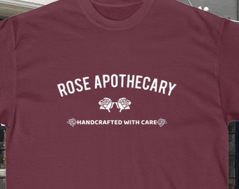 May include: A maroon t-shirt with white text that reads "ROSE APOTHECARY" and "HANDCRAFTED WITH CARE". The text is in an arched font with two stylized roses between the words.