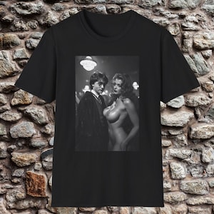 May include: Black t-shirt featuring a black and white image of two people. One person is wearing glasses and a dark robe, and the other is nude. The image is set against a stone wall background.