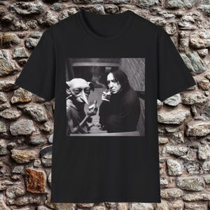 May include: Black t-shirt featuring a black and white image of two characters. One character is holding a cigarette, and the other is holding a glass. The image is set against a brick wall background.