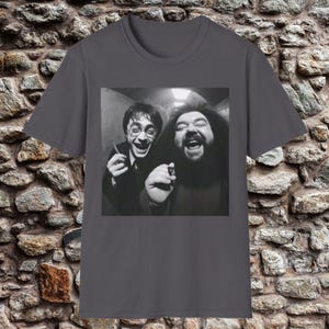 May include: Dark gray t-shirt featuring a black and white image of two people laughing. One person wears glasses and holds a small object. The other person has a long beard and also holds a small object. The image is centered on the shirt.