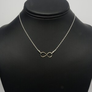 May include: A silver-toned necklace with an infinity symbol pendant. The chain is delicate, and the infinity symbol is centered. The necklace is displayed on a black mannequin, showcasing the jewelry's design and style.