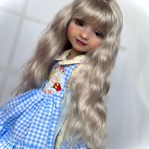 Könnte beinhalten: Eine Puppe mit langem, welligem, hellgrauem Haar und einem lächelnden Gesichtsausdruck. Die Puppe trägt ein blau-weißes Vichy-Kleid mit floralen und Cartoon-Akzenten am Oberteil. Die Haut der Puppe hat ein realistisches, bemaltes Aussehen.