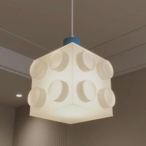 Op de afbeelding: Een witte, kubusvormige hanglamp met een blauwe basis. De lamp heeft cirkelvormige, verhoogde details aan elke kant, die op een bouwsteen lijken. Hij hangt aan een wit snoer.