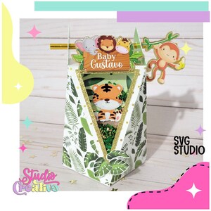 Peut inclure: Une boîte à dragées de fête sur le thème de la jungle, blanche et verte, avec une fenêtre transparente. La boîte est ornée d'une bordure dorée pailletée et représente un tigre, un singe, un éléphant et un lion. La boîte porte une pancarte indiquant "Baby Gustavo".
