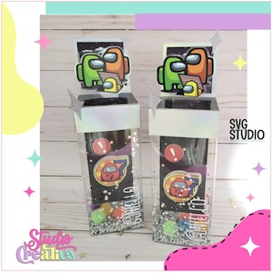 Puede incluir: Dos cajas de plástico transparente con un fondo de espacio negro y blanco. Las cajas tienen un borde de purpurina plateada y contienen pequeñas bolas de colores. La parte superior de cada caja tiene una imagen impresa de personajes del videojuego Among Us. El texto "Game On" está impreso en la parte delantera de cada caja.