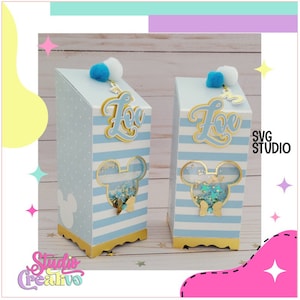 Puede incluir: Dos cajas rayadas en azul y blanco con ribetes dorados y un recorte de cabeza de Mickey Mouse. Cada caja tiene un pompón blanco en la parte superior y una etiqueta de nombre dorada que dice "Zoe". Las cajas están llenas de purpurina y tienen una ventana transparente.