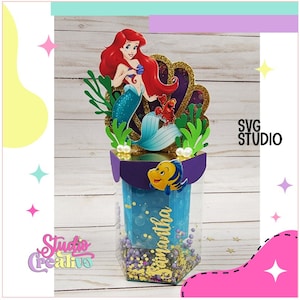 Puede incluir: Una caja de plástico transparente llena de confeti dorado y morado, con un fondo azul y el nombre "Samantha" escrito en ella. La caja tiene una tapa morada y dorada con una figura de sirena en la parte superior. La sirena tiene el pelo rojo y lleva una cola verde. La figura sostiene una concha y un cangrejo.