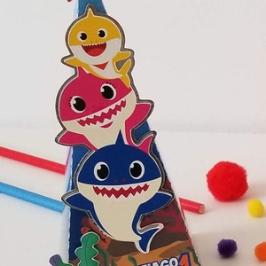 Puede incluir: Un cono de fiesta colorido con un dise&ntilde;o de tibur&oacute;n azul, amarillo y rosa. El cono tiene una estrella roja en la parte superior y las palabras "Santiago 1 Cumple" en el lateral.
