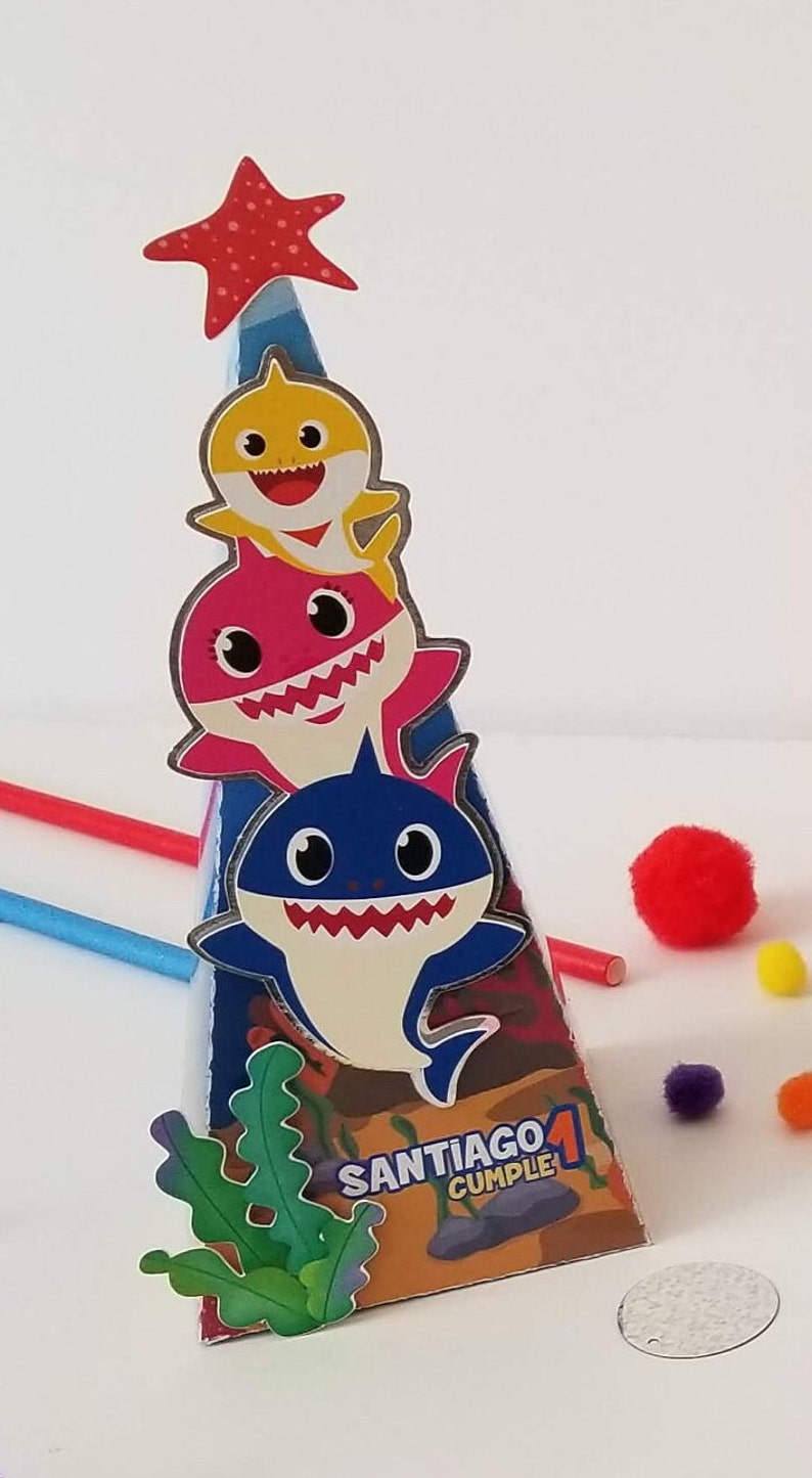 Puede incluir: Un cono de fiesta colorido con un dise&ntilde;o de tibur&oacute;n azul, amarillo y rosa. El cono tiene una estrella roja en la parte superior y las palabras "Santiago 1 Cumple" en el lateral.