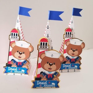 Puede incluir: Tres cajas de regalo en forma de cono decoradas con un tema n&aacute;utico. Cada caja presenta un osito de peluche con un gorro de marinero y una camisa a rayas rojas y blancas. Las cajas est&aacute;n coronadas con una bandera azul y un dise&ntilde;o de faro. El texto "Daniel Alejandro" est&aacute; impreso en cada caja.