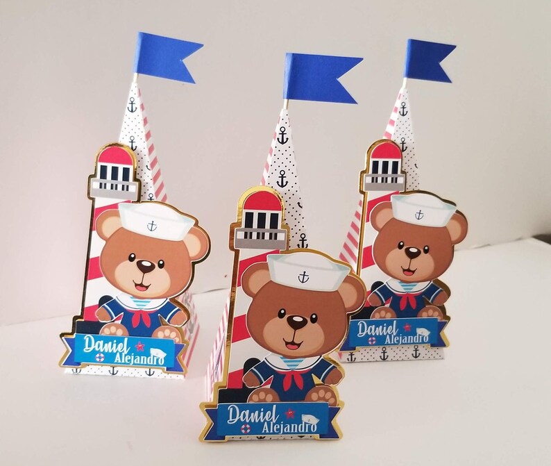 Puede incluir: Tres cajas de regalo en forma de cono decoradas con un tema n&aacute;utico. Cada caja presenta un osito de peluche con un gorro de marinero y una camisa a rayas rojas y blancas. Las cajas est&aacute;n coronadas con una bandera azul y un dise&ntilde;o de faro. El texto "Daniel Alejandro" est&aacute; impreso en cada caja.