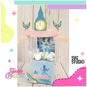 Puede incluir: Una caja de favor de fiesta de temática de Cenicienta azul y dorada con una ventana de plástico transparente. La caja está decorada con una torre de reloj, pájaros y una pancarta rosa. La caja está llena de chocolates envueltos en papel plateado y tiene una zapatilla de Cenicienta en la parte inferior. La caja dice "Anabella 3 cumple".