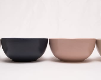 May include: Three ceramic bowls in different colours: dark grey, light pink, and beige. The bowls are lined up in a row on a white surface.