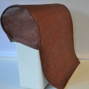 May include: A brown, curved leather headrest cover. The cover has a textured appearance and a deep brown hue. The design is a rounded shape, suggesting it is designed to fit over a chair or seat back. The headrest cover is a solid colour.