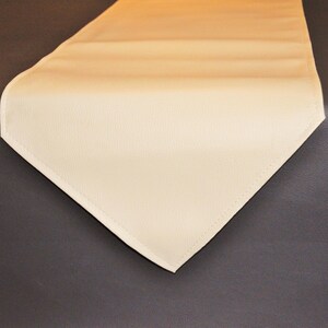 May include: A cream-coloured table runner with a subtle texture. The runner is made of a soft, smooth material and has a slightly rounded edge.