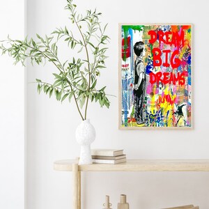K&ouml;nnte beinhalten: Ein farbenfroher abstrakter Kunstdruck mit einer jungen Person, die vor einer Backsteinmauer steht. Der Druck zeigt die Worte "Dream Big Dreams" in roter Farbe.